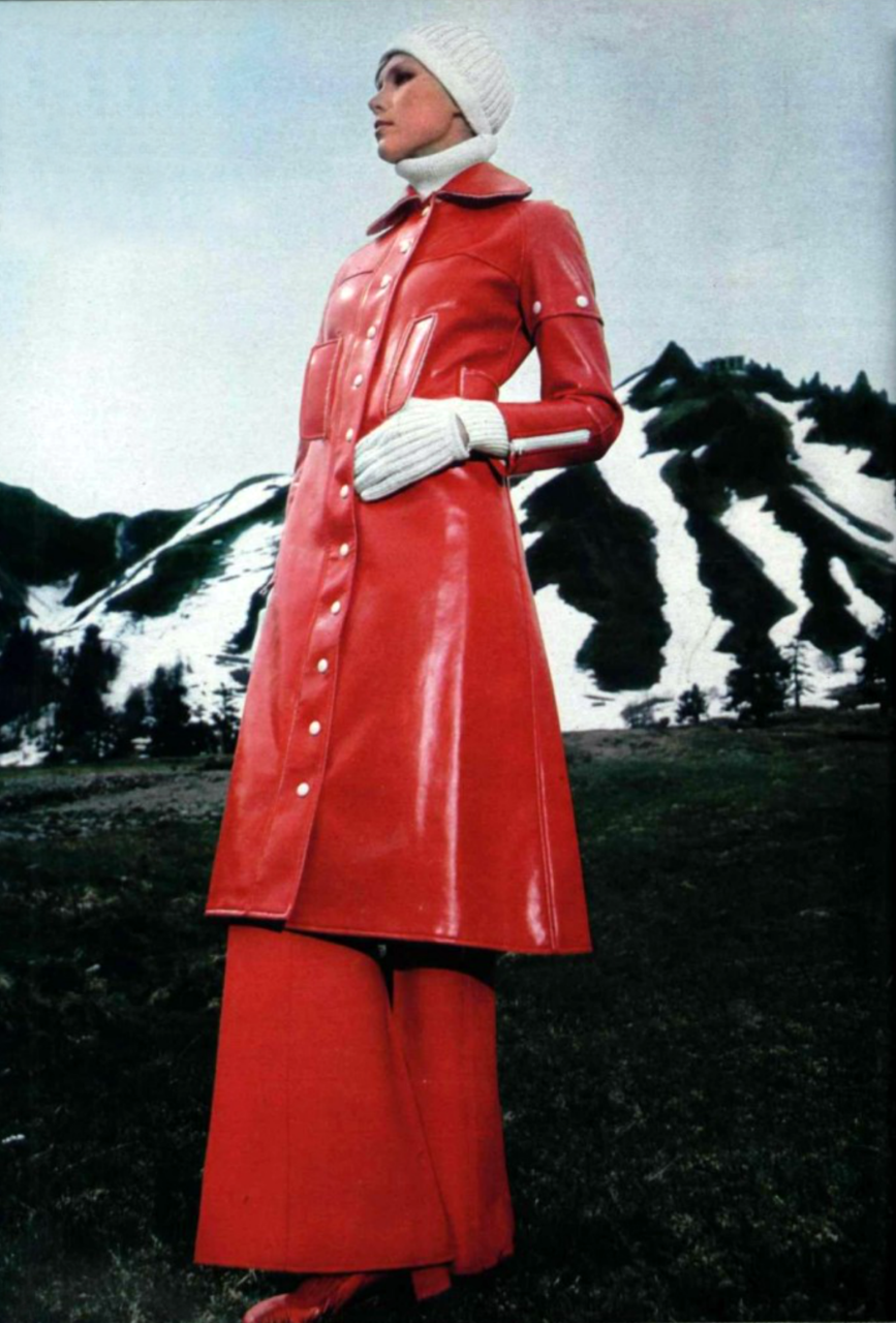 Fashion model in a glossy red coat and pants, white knit hat and gloves, posing against snowy mountains.