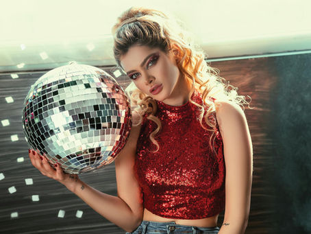 Woman in red sequined top holds a disco ball, surrounded by light reflections. Her expression is contemplative, against a modern interior.