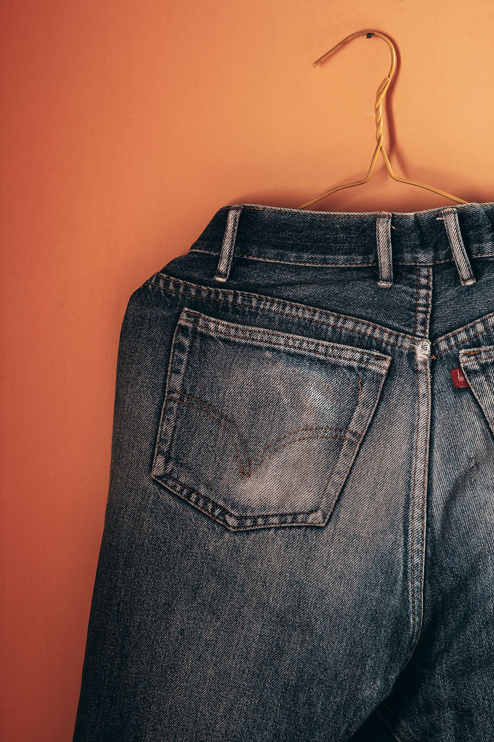 Vintage Levi jeans hanging on a gold hanger against a warm orange wall, with a single back pocket and casual retail-style mood