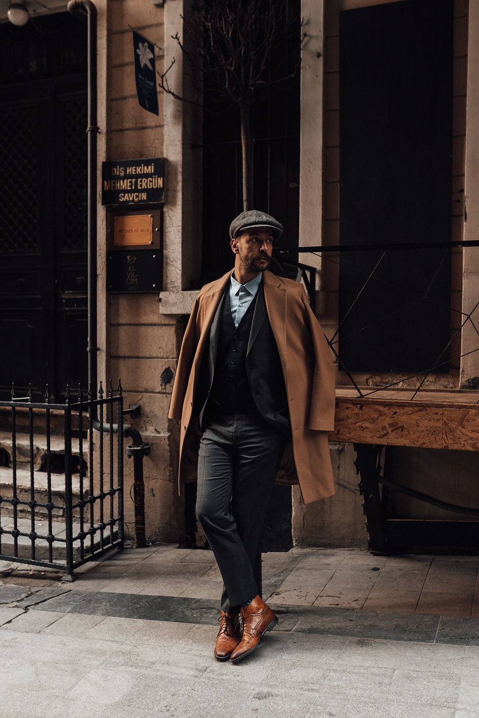 Peaky Blinders vibes. Stylish man in tan coat, vintage suit and flat cap leans outside a stone building with Turkish signs, looking pensive on a quiet street.