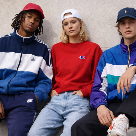 Three people in colorful sporty jackets sit casually against a concrete wall. They wear caps and display logos like Nike and Champion.