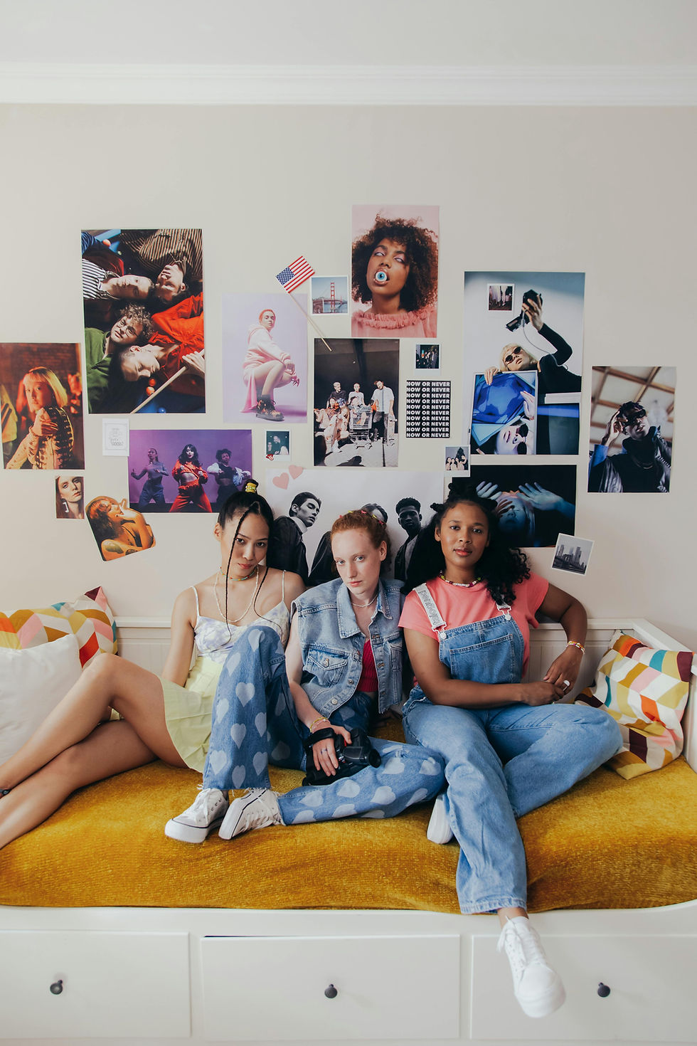 90s aesthetic: Three friends sit on a bed with colourful cushions. Walls are covered in eclectic posters. Mood is relaxed and casual.