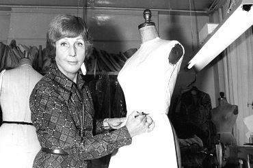 Black and white photo of Maureen Baker in a dressmaking studio sketches on a white dress form, with mannequins and fabric in the background.