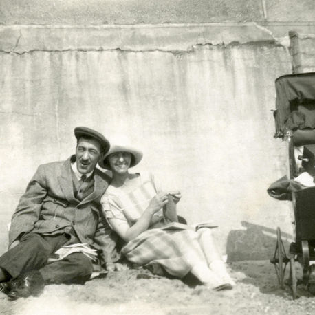1920s black and white photograph. A man and woman sit on the sand, smiling. The woman knits; next to them is a vintage stroller. They're against a textured wall. Mood is relaxed.
