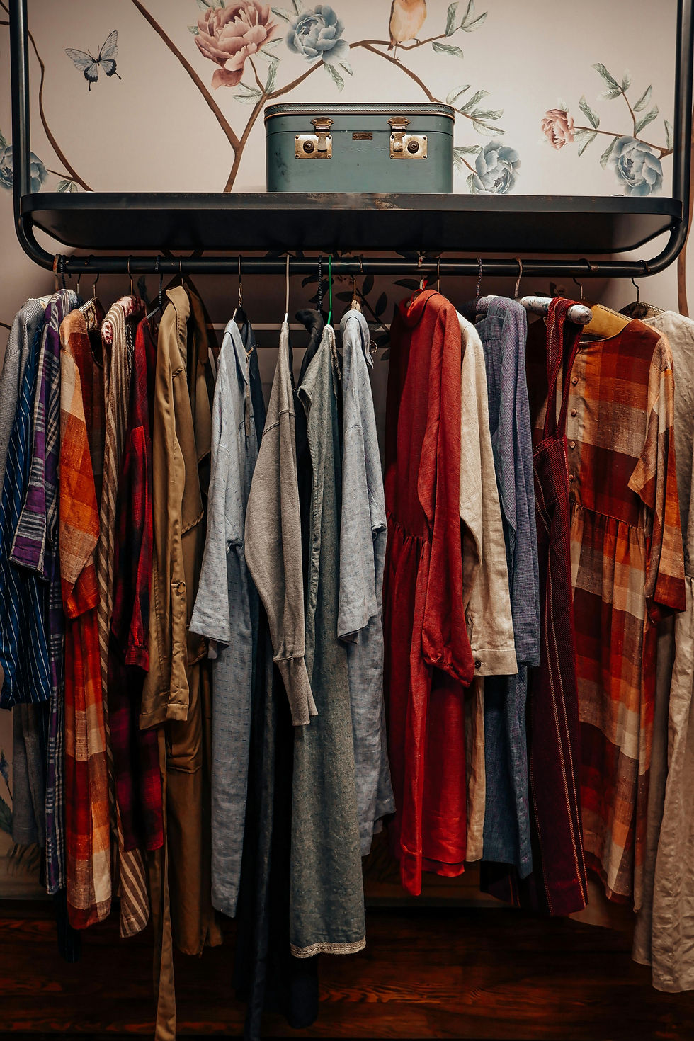Clothes in various colors and patterns hang in a closet. A vintage suitcase sits on a shelf above. Floral-patterned wallpaper is in the background.