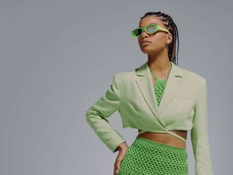 Fashion model in neon green sunglasses, green blazer and green knit skirt posing against a plain grey studio backdrop.