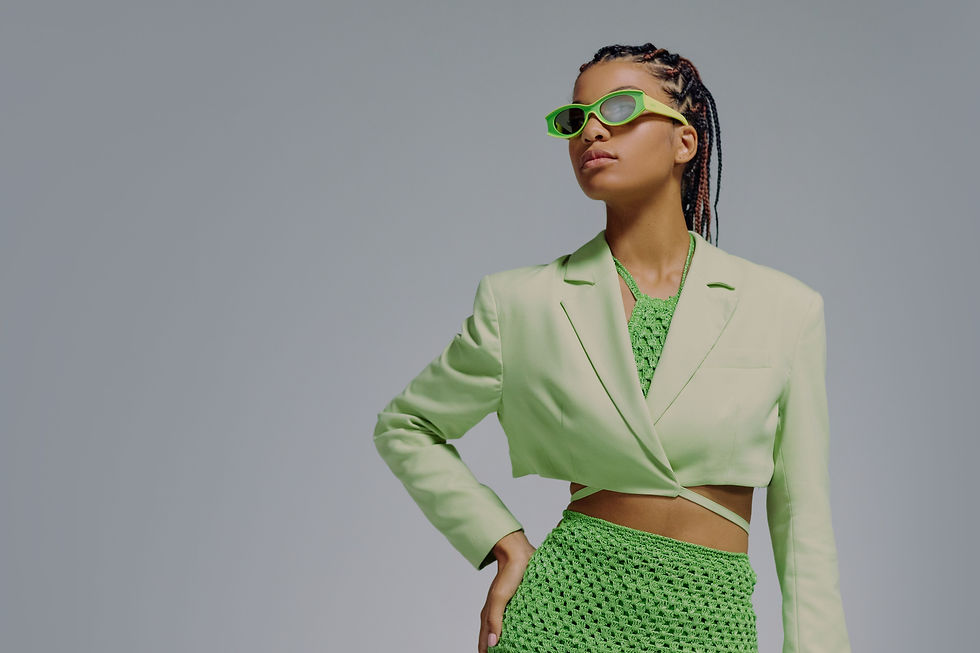 Fashion model in neon green sunglasses, green blazer and green knit skirt posing against a plain grey studio backdrop.