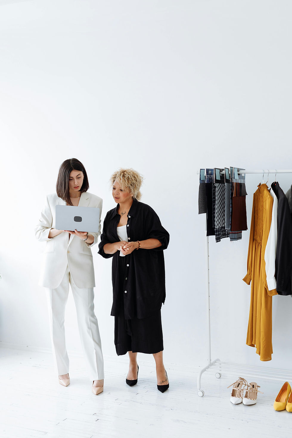 Two women review a laptop in a bright white fashion studio beside a rack of colourful clothes and shoes