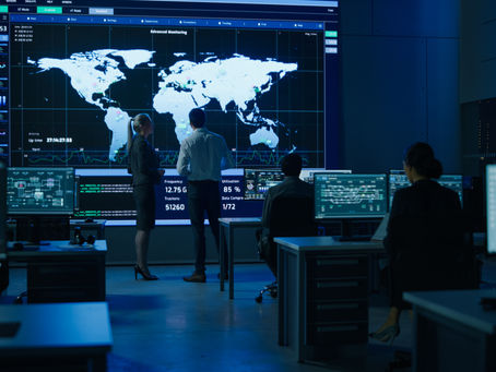 People analyze a large world map display in a dim control room with multiple monitors, showing data and graphs. Atmosphere is high-tech.