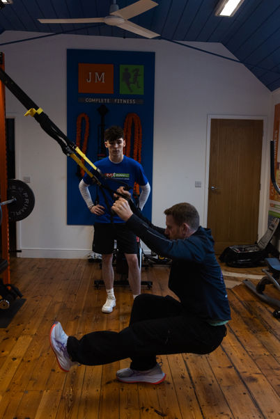 John, demonstrating a TRX pistol squat to a motorsport athlete in the JM Complete Fitness studio as part of an elite strength and conditioning session.