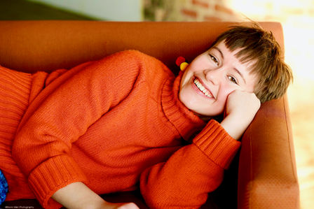 Senior picture of a young woman laying on her side on an orange couch wearing an orange sweater. Photo by Bonni Allen.