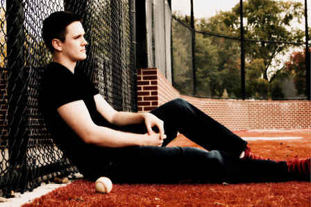 Baseball player senior picture inspiration featuring a young man sitting against a chain link fence with a baseball in the foreground. Photo credit Bonni Allen Photography.