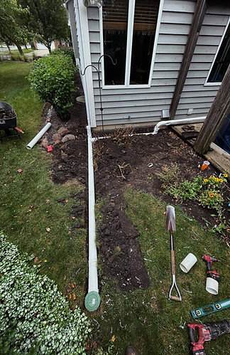Downspout drainage pipe installation in progress along the side of a gray suburban home, with white PVC pipe running from the foundation through a freshly dug trench to a green cleanout cap in the yard