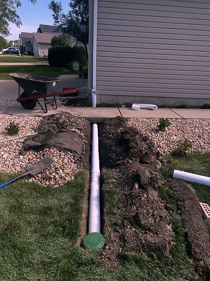 French drain installation in progress alongside a residential home in Joliet, IL, showing drainage pipe laid in a trench with excavated soil and rock bed