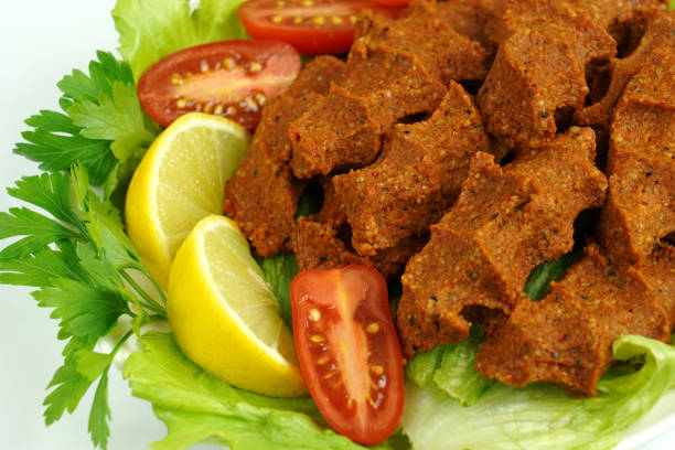 Yeşillik ve limon dilimleri ile sunulmuş çiğ köfte tabakta. Yanında kesilmiş domatesler. Yemek iştah açıcı görünüyor.