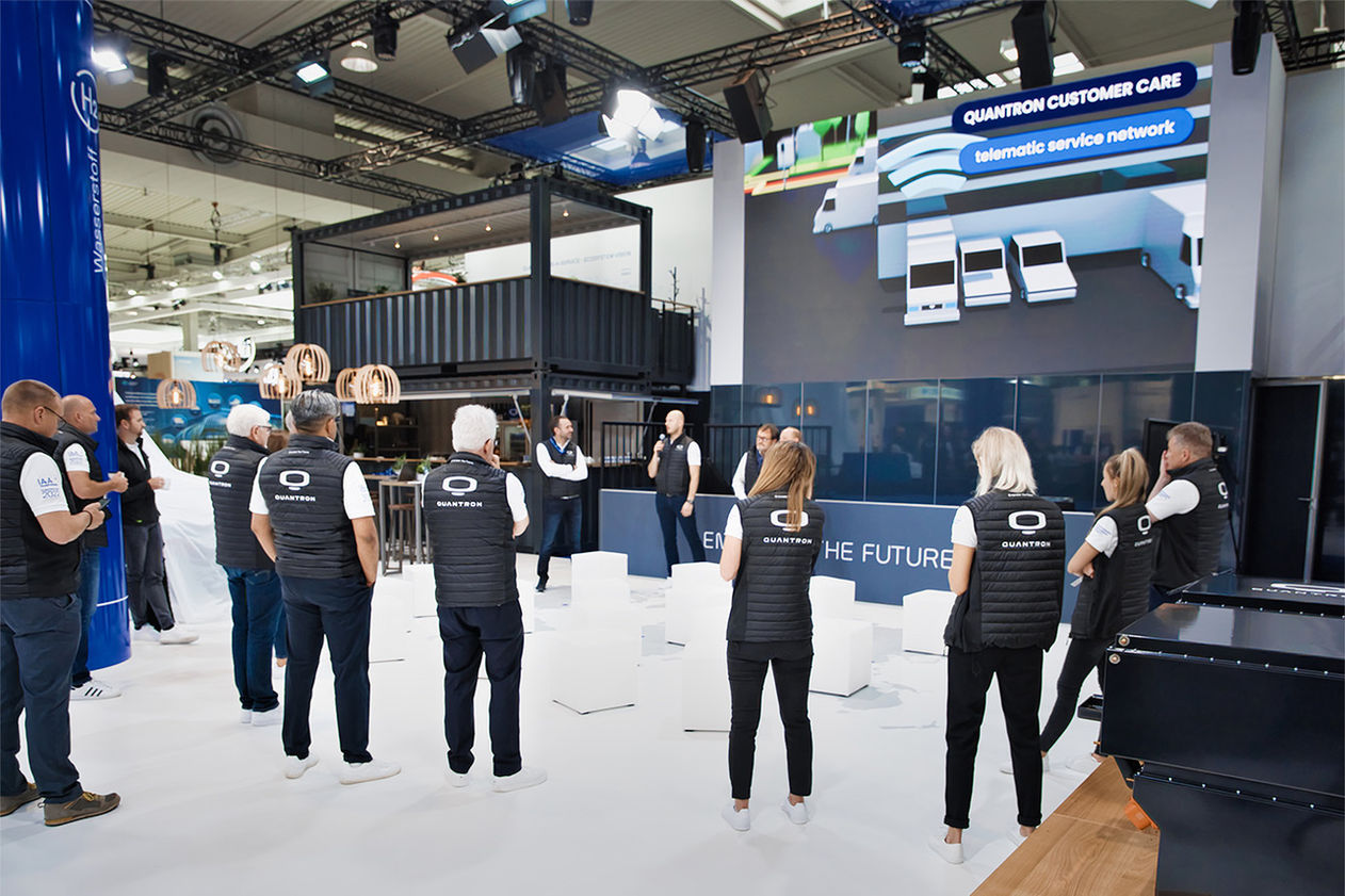 Qunatron Team steht auf dem Messtand und hören einer Rede zu.