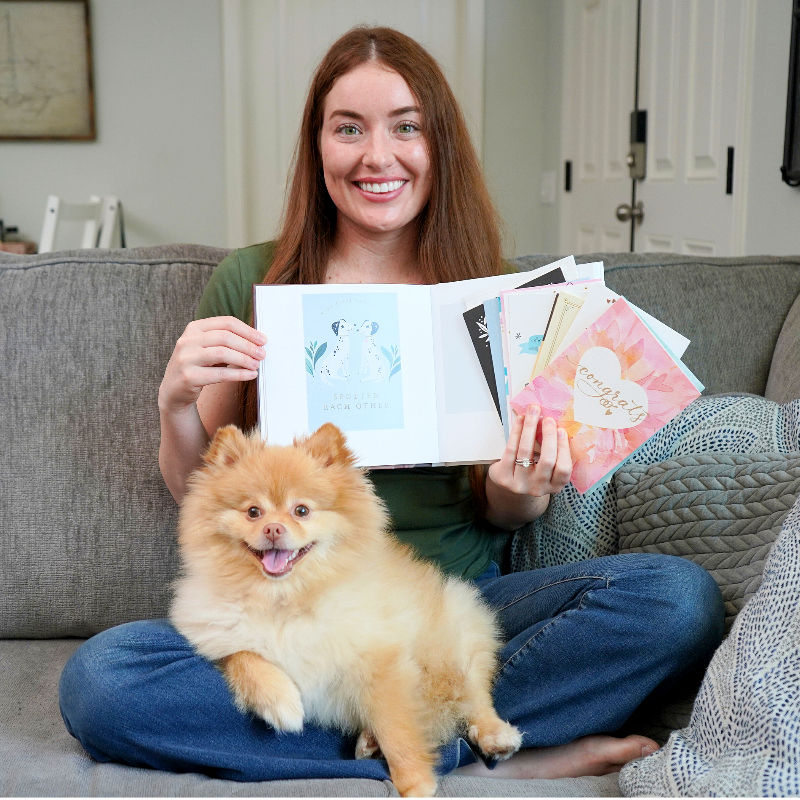 A Bride-to-be holds an open a bridal shower memory book filled with greeting cards, with her Pomeranian in her lap