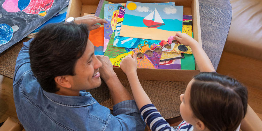 A dad and his young child looking through a box of saved kids' artwork before sending it in for a memory book