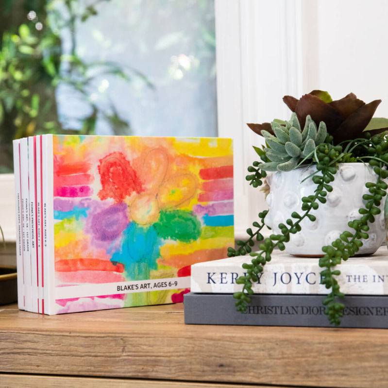 A stack of Artkive memory books displayed on a mantle next to a decorative plant, keepsake books made from children's artwork