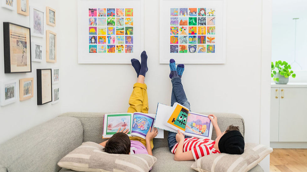 Two children relax on a couch with their feet on the wall next to Artkive mosaics. Together, they look over their art books. (art of scribbles, encouraging creativity, young children)