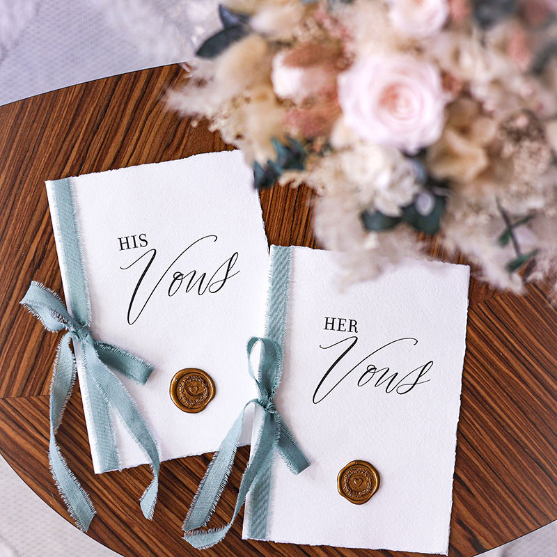 Two handwritten wedding vow booklets labeled "His Vows" and "Her Vows," tied with ribbon and wax seals beside a bouquet