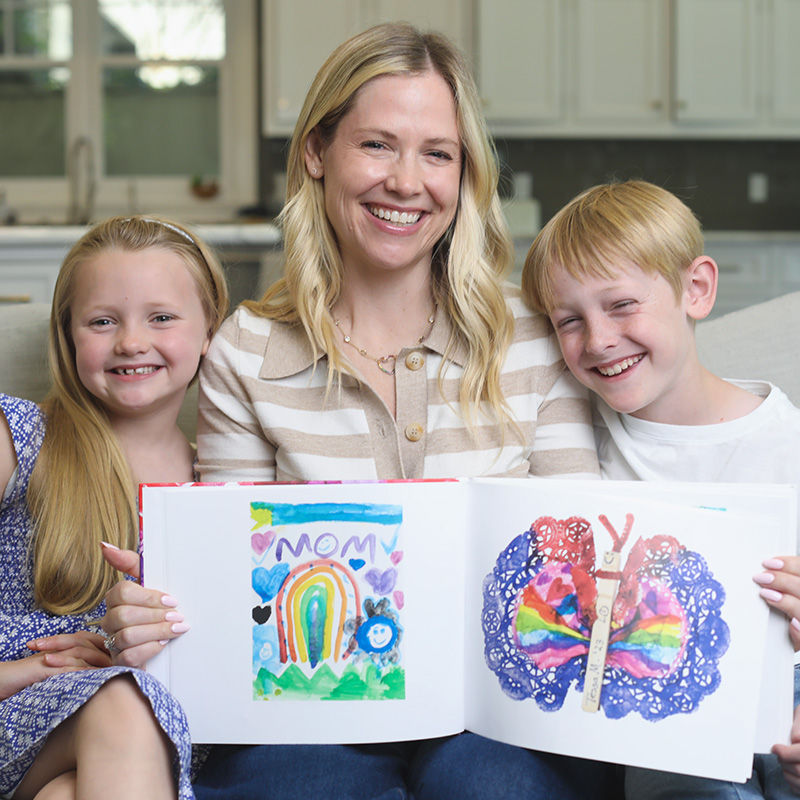 Mom sits proudly with her two children, holding an open Artkive kids' art memory book together at home.