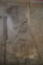 Ancient stone relief of a bearded man sitting on a throne, holding a staff and flower. Detailed carvings with an ornate chair design.
