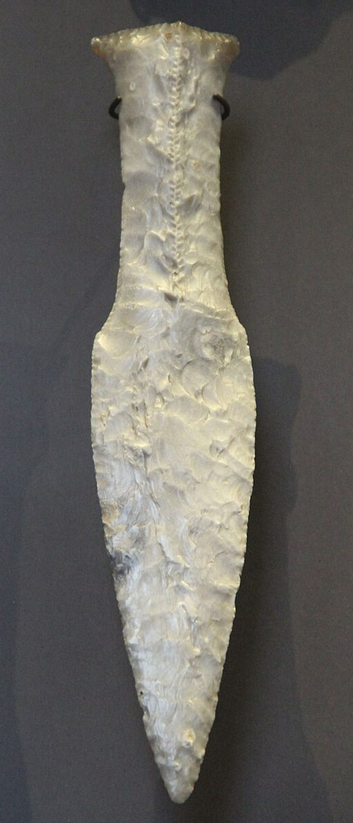 A flint knife with a textured surface and pointed tip, set against a plain dark background. The blade appears ancient and handcrafted.