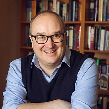 Smiling man in glasses and a sweater vest sits in front of a bookshelf filled with books, creating a warm, intellectual atmosphere.