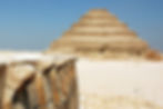 Ancient step pyramid in a desert with a clear blue sky, foreground features stone carvings of cobras on a ledge, evoking a historical mood.