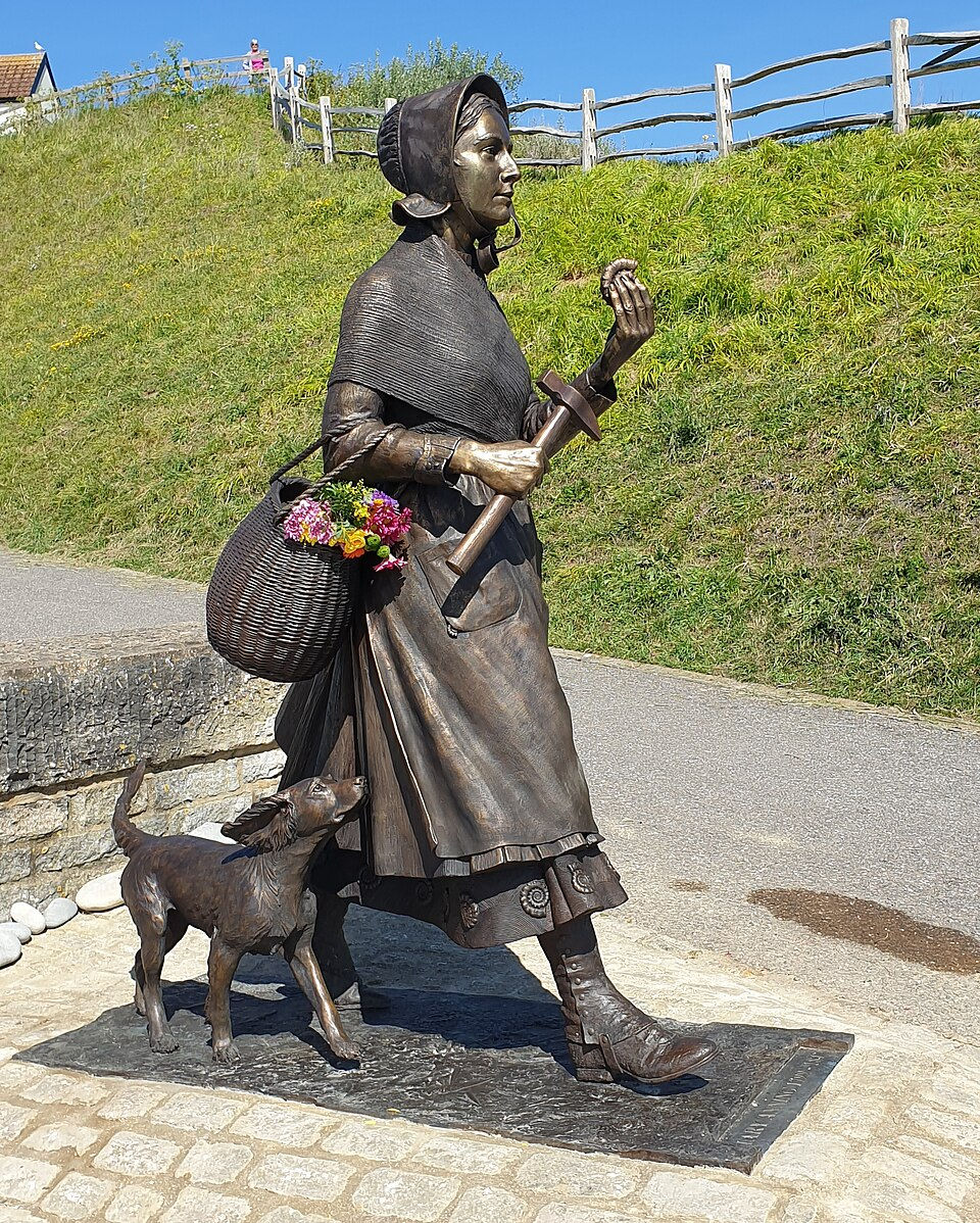 Bronze statue of a bonneted woman walking with a basket of flowers and a dog beside her on a sunny grassy hillside path.