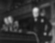 Winston Churchill speaking at a podium with CBC microphones, dressed in a suit and bowtie, in a formal setting with a wood-paneled background.