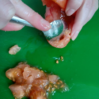 The pulp of a tomato is being scraped out by a spoon. Pulp from the tomato is in a pile on a green mat nearby.