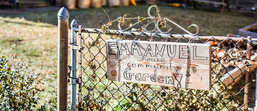 A wooden sign on a wire fence says "Emmanuel House Community Garden."