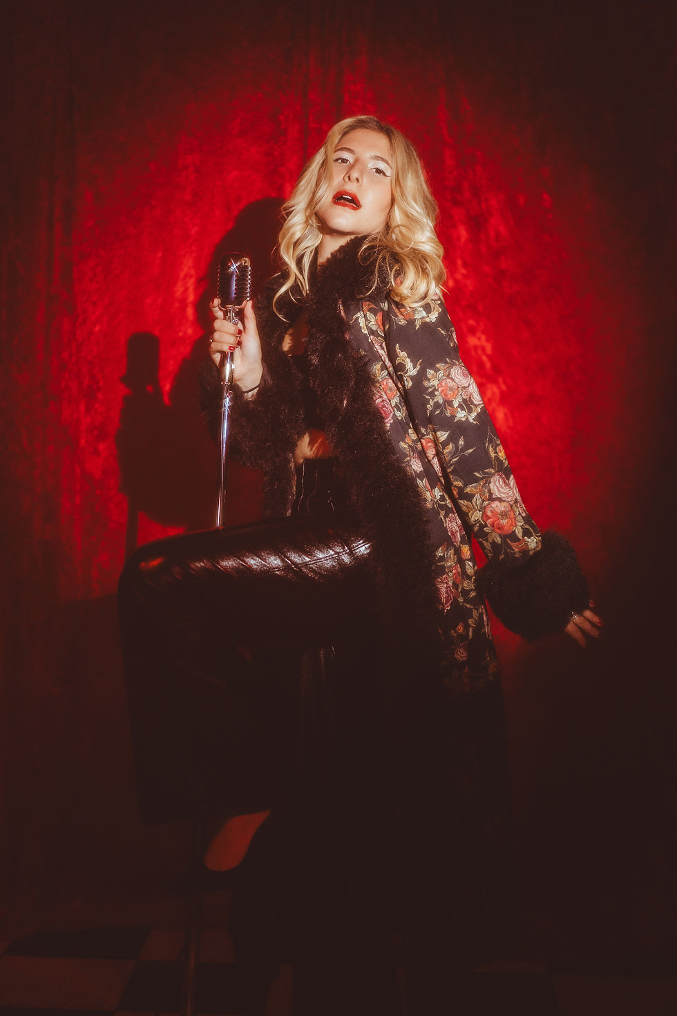 Singer in a floral coat poses with vintage mic against a red backdrop, exuding a dramatic and glamorous vibe. Goldie Scott - The Cage, a music blog powered by Cage Riot