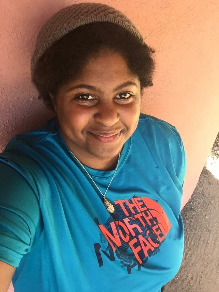 Smiling person wearing a blue The North Face shirt and beanie, leaning against a pink wall in a sunny setting. Relaxed and cheerful mood. Elena C. Lockleis - The Cage, a music blog powered by Cage Riot