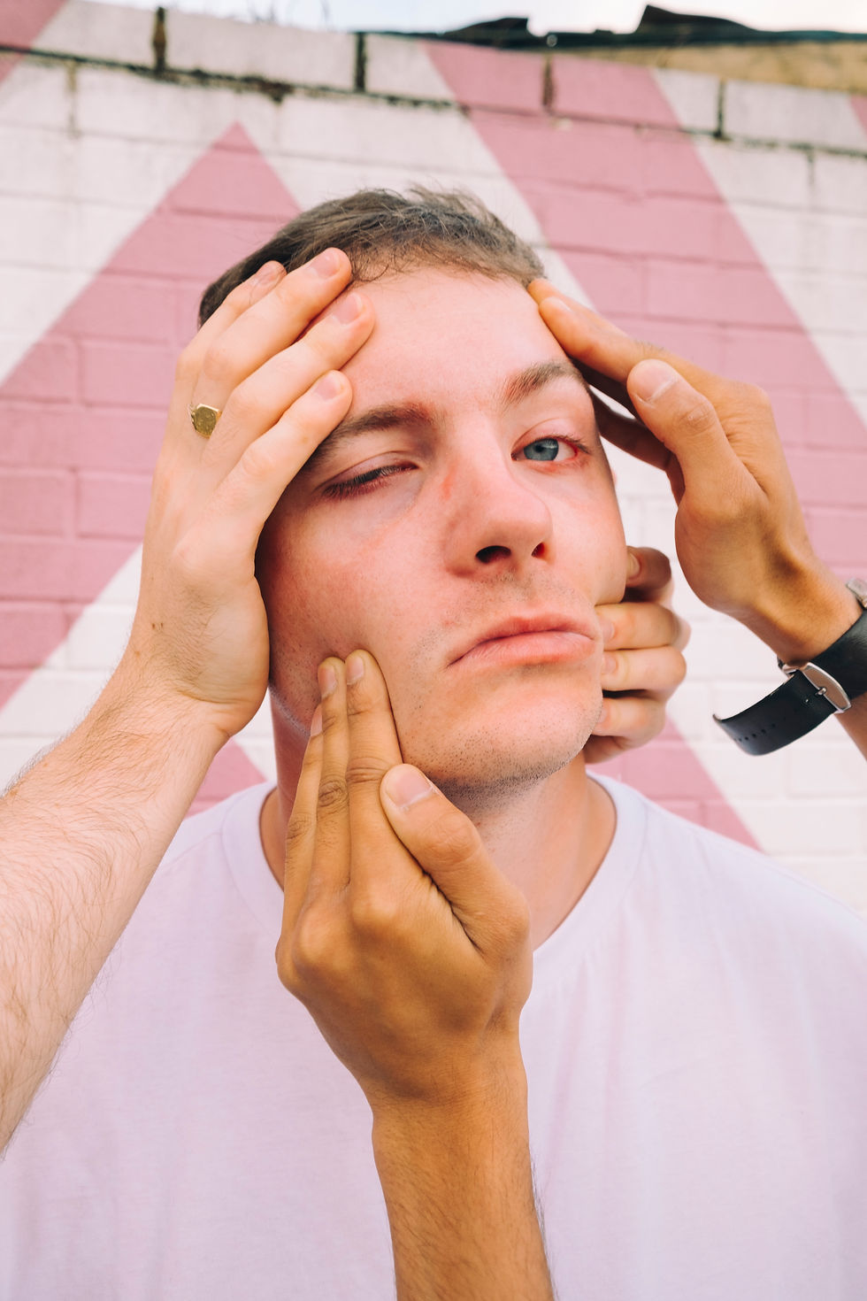 Man in white shirt with neutral expression, framed by hands against a pink and white brick wall. Hands adjusting face; calm mood. SPEAK! - The Cage, a music blog powered by Cage Riot