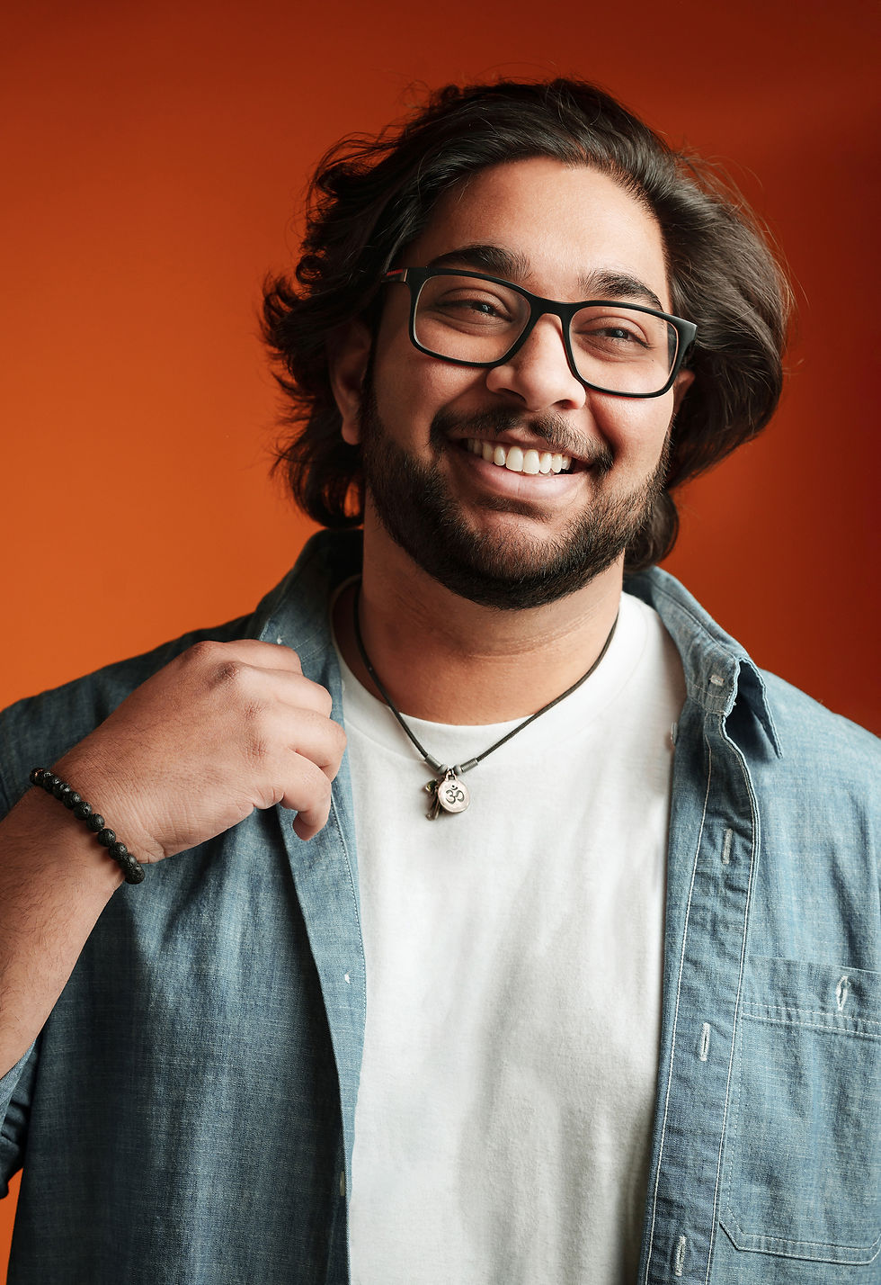 Smiling person with glasses wearing a necklace and denim shirt against an orange background, exuding a cheerful, relaxed vibe. Varun Sheel - The Cage, a music blog powered by Cage Riot