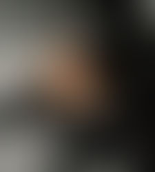 Young woman in black Adidas tracksuit, sitting on a gray sofa with a pensive expression. Dimly lit room with plant in background. Olivia Booth - The Cage, a music blog powered by Cage Riot