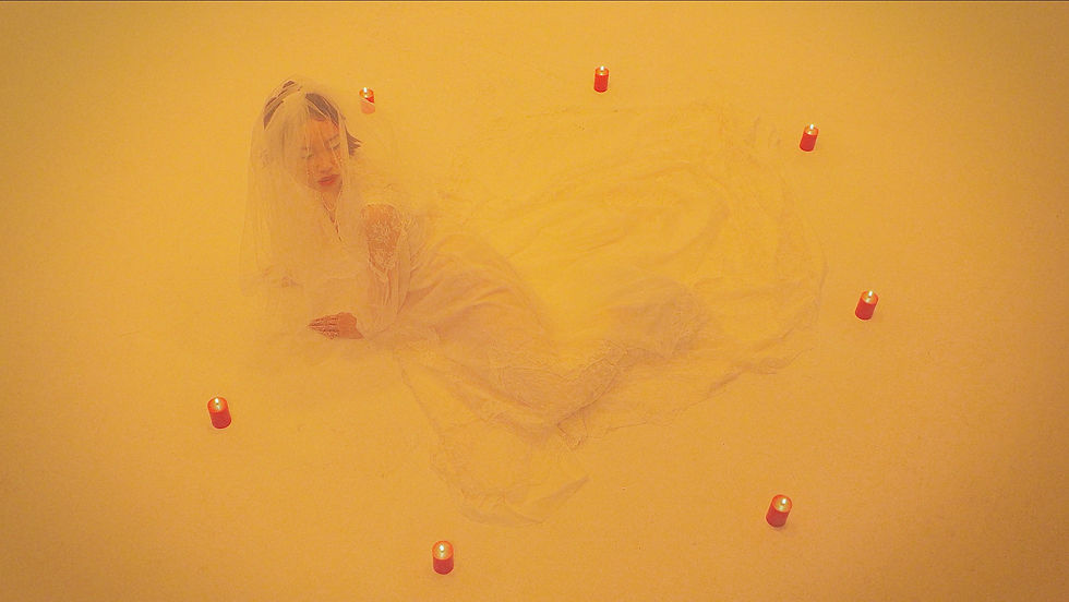 Bride in white dress lies on yellow background, surrounded by lit red candles, creating a serene, ritualistic atmosphere. Juliet By Night - The Cage, a music blog powered by Cage Riot