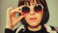 Woman with a bob haircut wears oversized pink sunglasses, adjusts them with one hand. She wears a black top and patterned jacket. Neutral background.