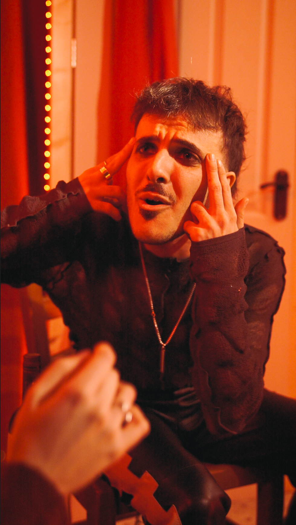 Man with expressive face gestures intensely, wearing black top, necklace; sitting indoors with red lighting and curtains creating a dramatic mood. Ren Shelter - The Cage, a music blog powered by Cage Riot