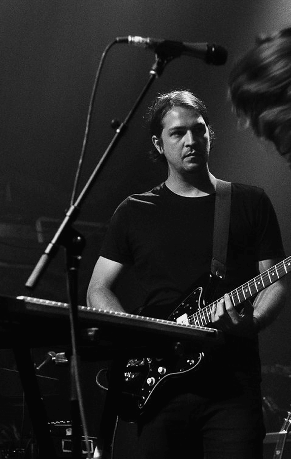 Musician in black shirt plays electric guitar on stage under dim lighting, focused expression. Microphone and another musician partially visible. Tropigloom - The Cage, a music blog powered by Cage Riot