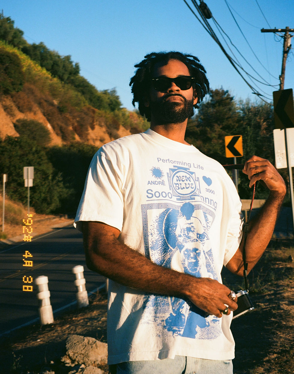Man in sunglasses holding a camera on a sunny road. Wearing a white T-shirt with blue designs. Trees and road signs in the background. Noah Kahlil - The Cage, a music blog powered by Cage Riot