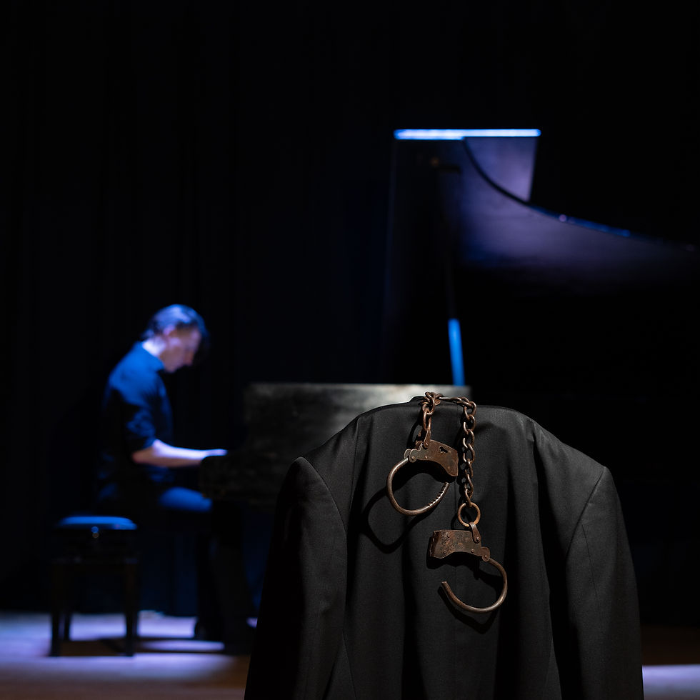 A person plays piano under a spotlight. A black jacket with handcuffs draped over it is in the foreground, set against a dark background. Micah Rose-Trespeuch - The Cage, a music blog powered by Cage Riot