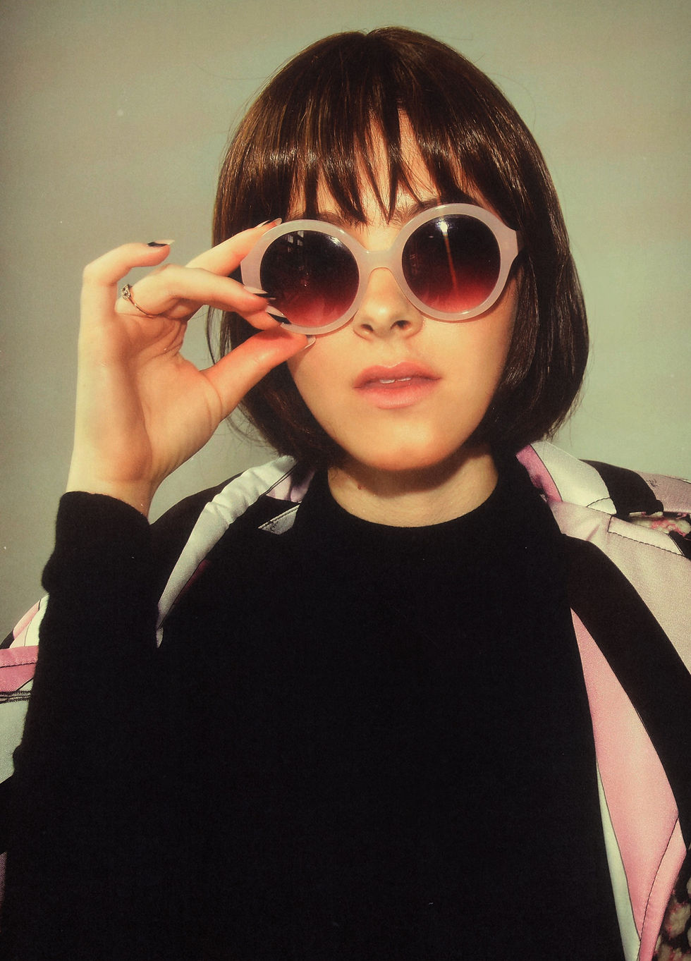 Woman with a bob haircut wears oversized pink sunglasses, adjusts them with one hand. She wears a black top and patterned jacket. Neutral background.