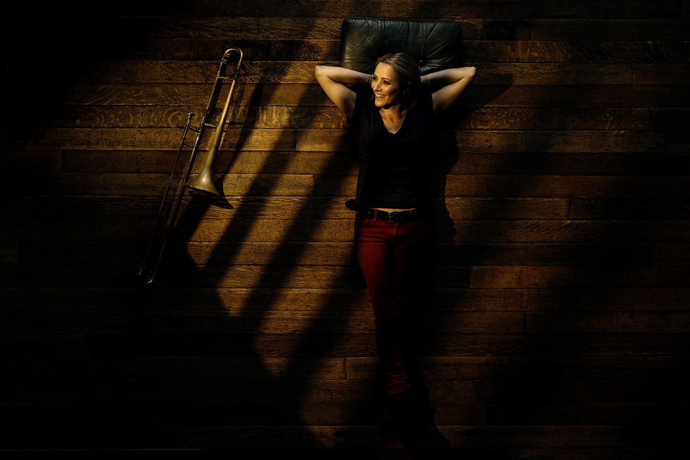 Woman in red pants lies relaxed on wood floor with a trombone nearby, surrounded by soft, warm light and shadows. Carol Jarvis - The Cage, a music blog powered by Cage Riot