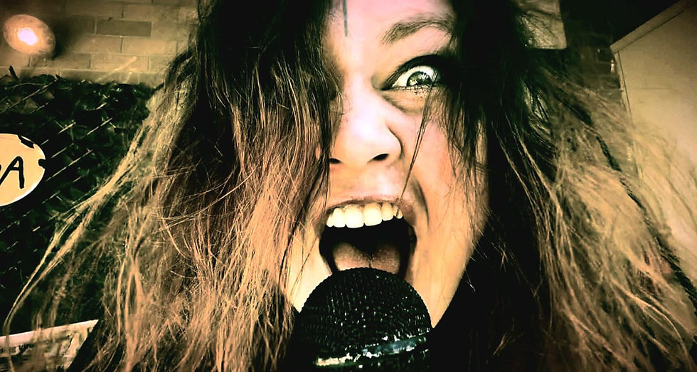 Close-up of a person with wild hair, holding a microphone, screaming intensely. Background shows a chain-link fence and partial sign. Clare Easdown- The Cage, a music blog powered by Cage Riot
