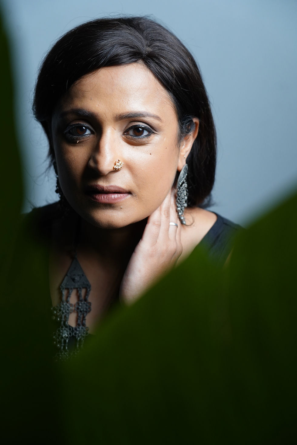 A woman with dark hair and jewelry poses, gazing forward. Soft focus greenery frames the image. The background is a plain blue-gray. Tarita - The Cage, a music blog powered by Cage Riot