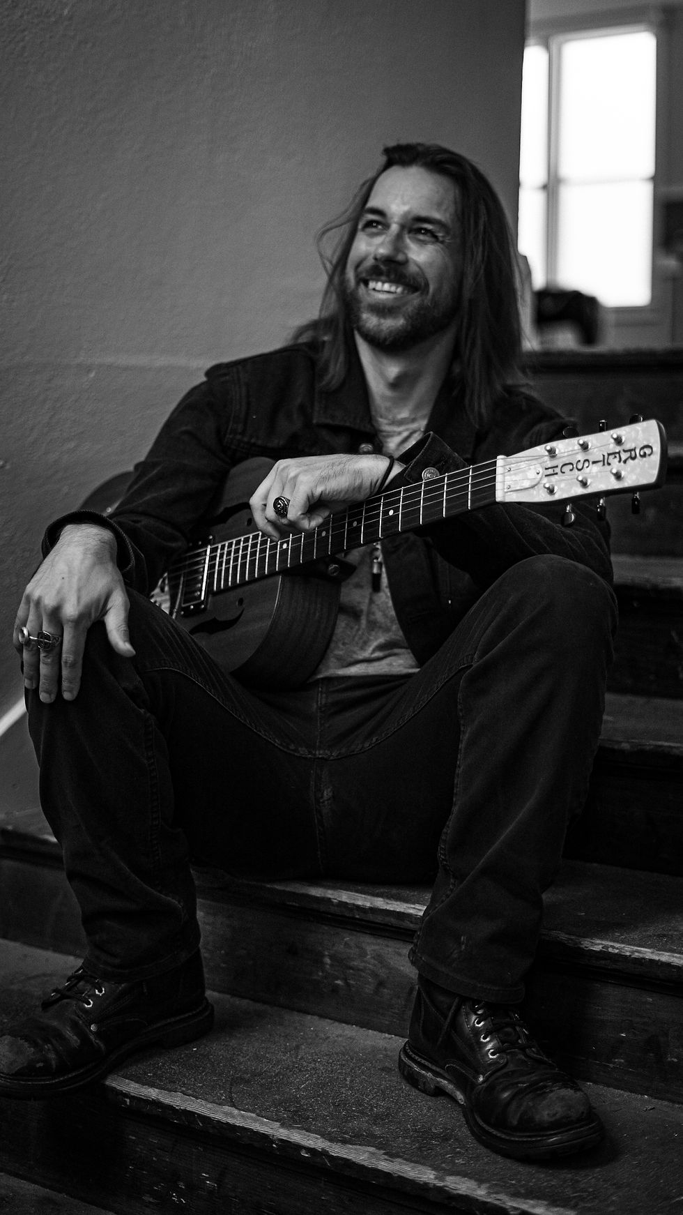 Man with long hair sitting on stairs, holding a guitar, smiling. He's wearing a jacket and boots. Black and white setting. Saint Nick the Lesser - The Cage, a music blog powered by Cage Riot